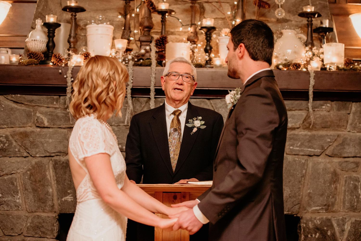 Bride and groom hold hands during indoor wedding ceremony before smiling officiant at candlelit stone fireplace.