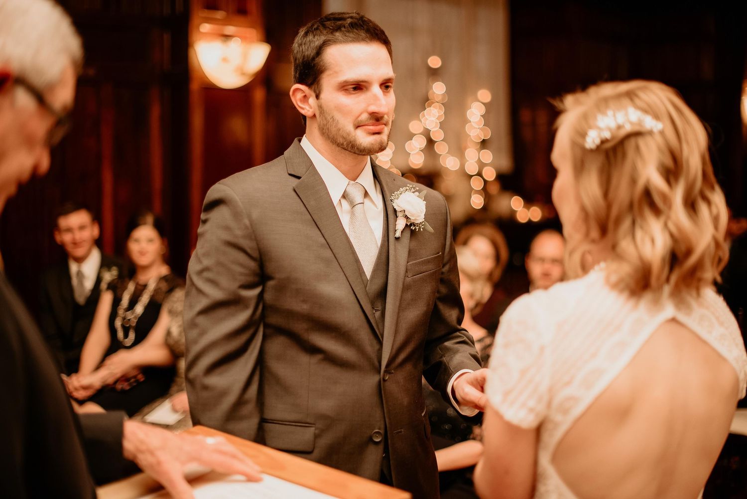 Groom in gray suit exchanges vows with bride in white dress during indoor wedding ceremony with warm bokeh lights.