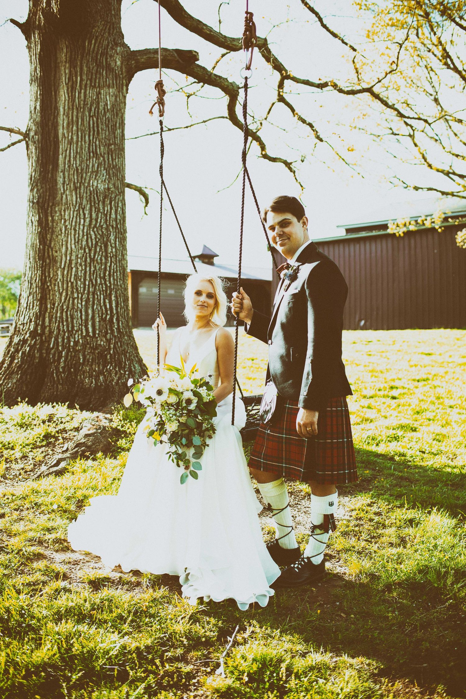 Bride in white gown holding bouquet stands with groom in red tartan kilt by a tree swing outdoors.