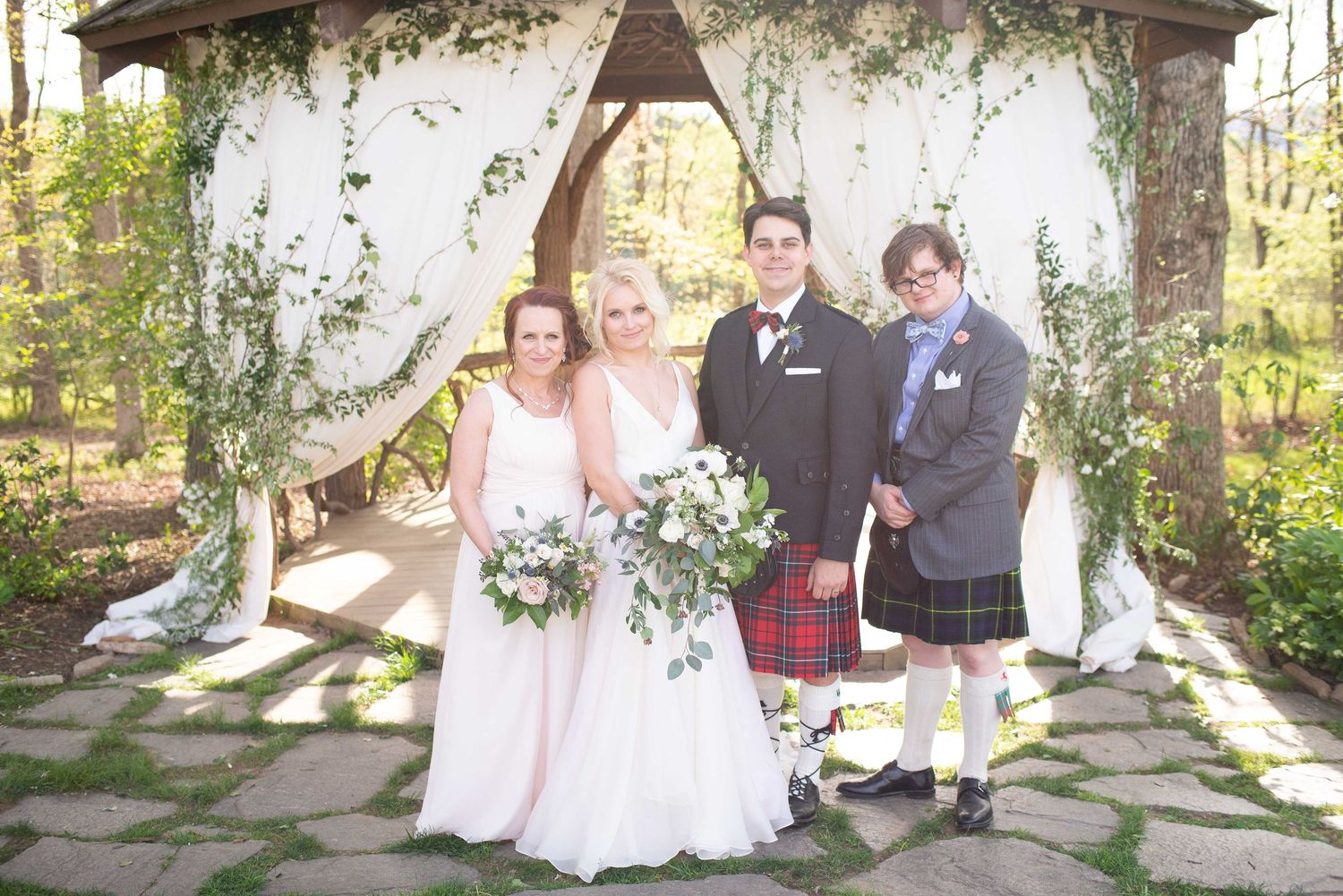 Wedding party poses under floral arch arbor outdoors, bride in white gown, guests in pink dress and Scottish kilt.