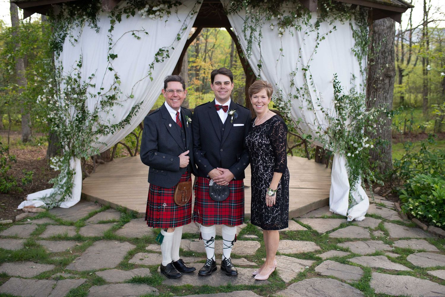 Groom in tartan kilt poses with parents under floral arbor at outdoor wedding ceremony.