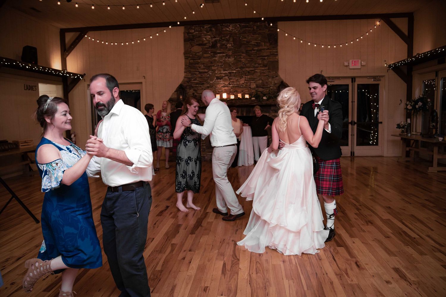 Couples dancing at a rustic wedding reception with string lights and a stone fireplace backdrop.