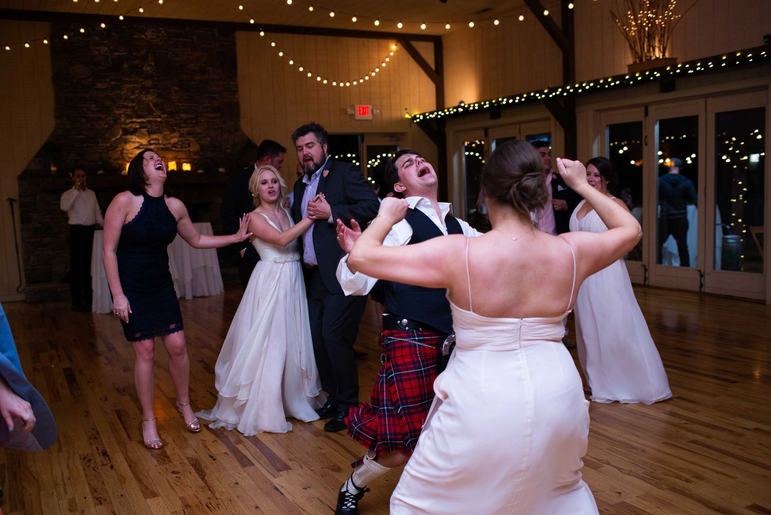 Wedding guests dancing joyfully at a reception in a rustic venue with string lights, featuring a groom in a kilt.