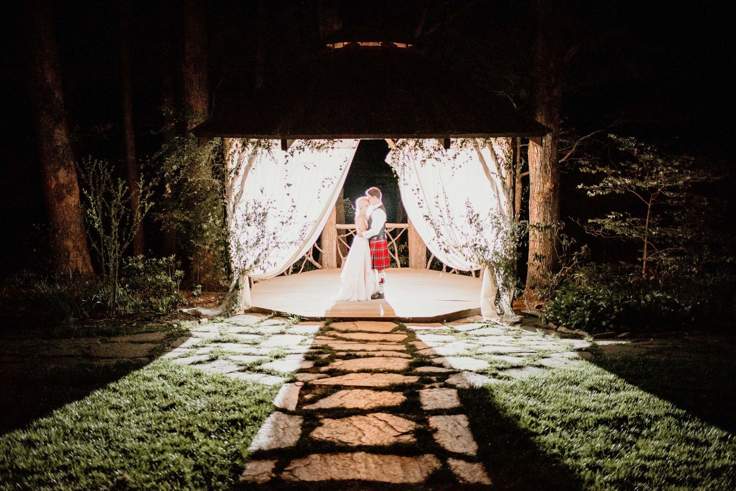 Couple sharing a romantic kiss under a glowing outdoor pavilion with white drapes and stone pathway at night.