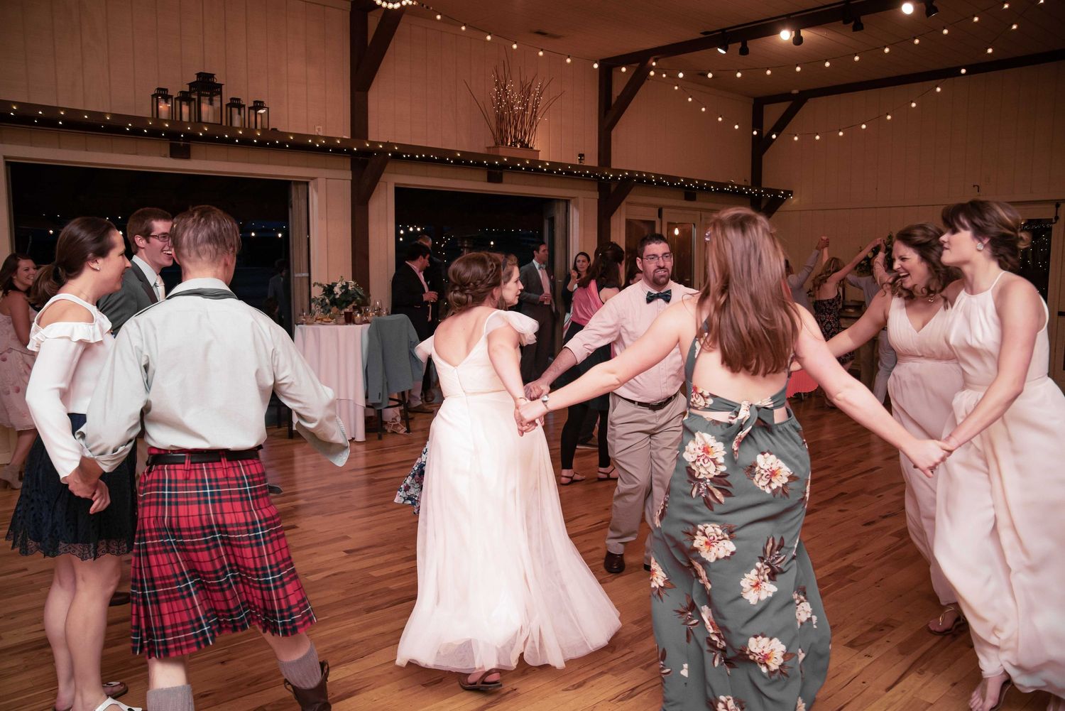 Wedding guests dancing in a circle on a wooden dance floor, including a man in a red tartan kilt, at a rustic barn reception.