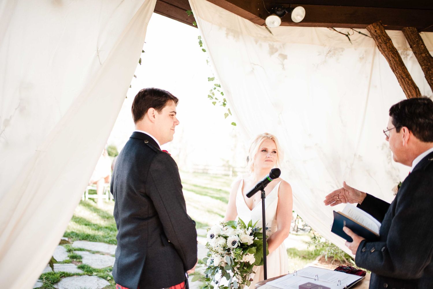 Bride reciting vows to groom at outdoor wedding ceremony under white draped canopy with officiant.