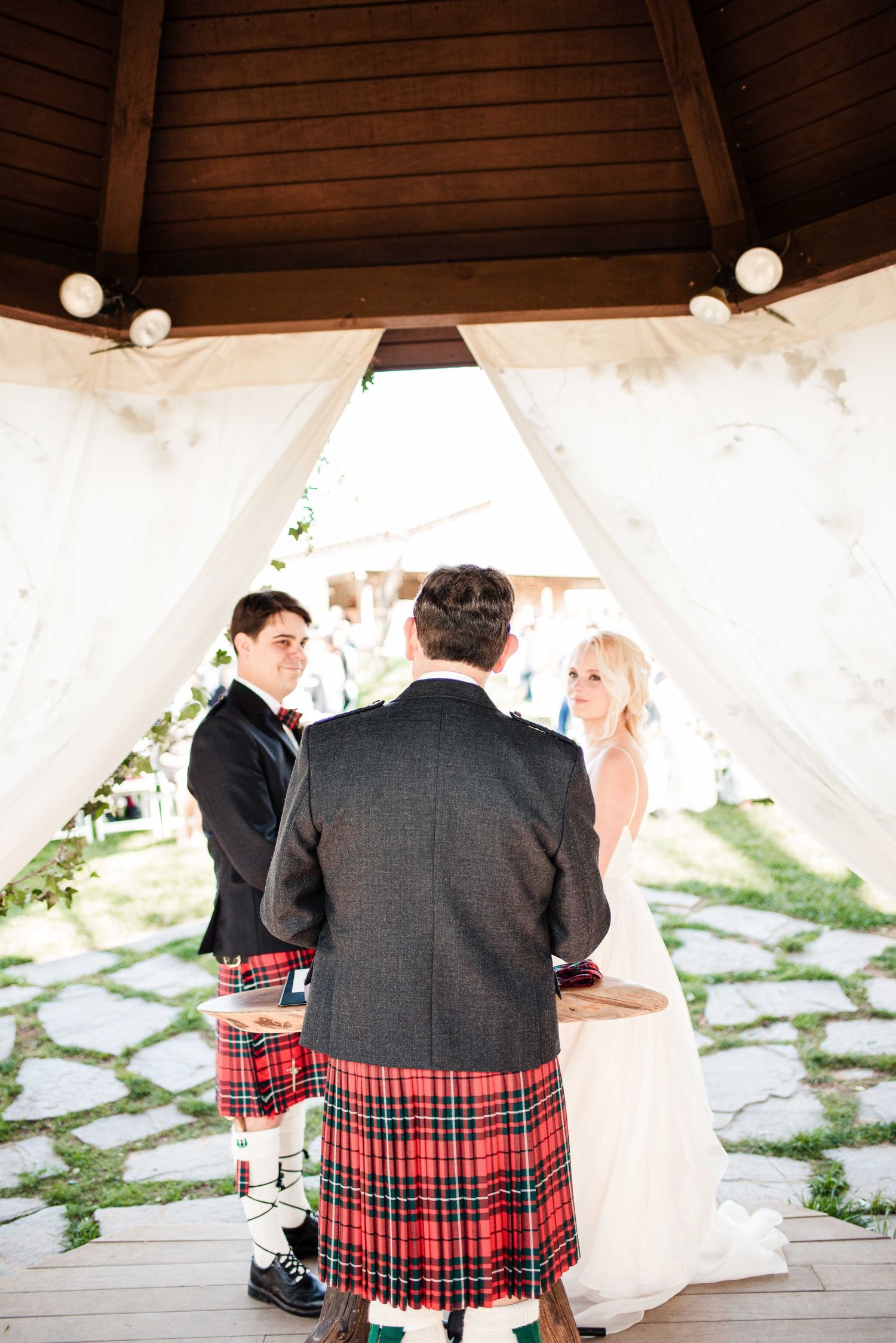 Wedding ceremony under gazebo with groom in red tartan kilt, bride in white gown, and officiant on stone path.