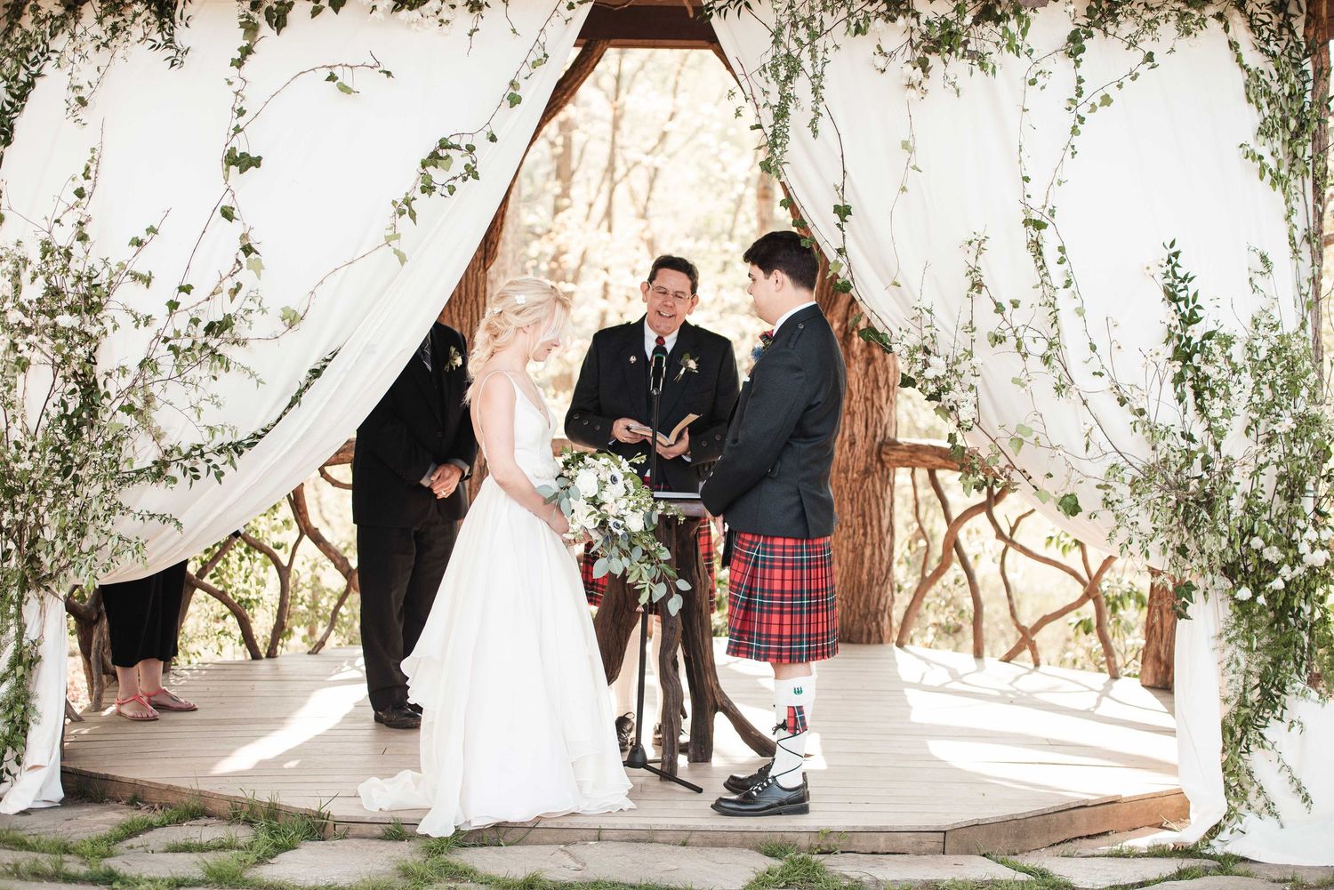 Bride and groom exchange vows under floral arch with officiant in kilt at outdoor woodland wedding ceremony.