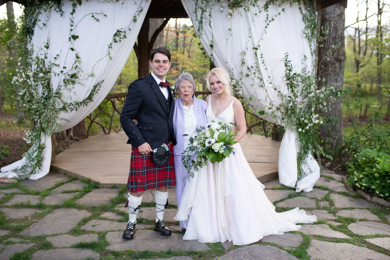 Bride and groom in Scottish kilt pose with elderly woman under floral arch at outdoor wedding ceremony.