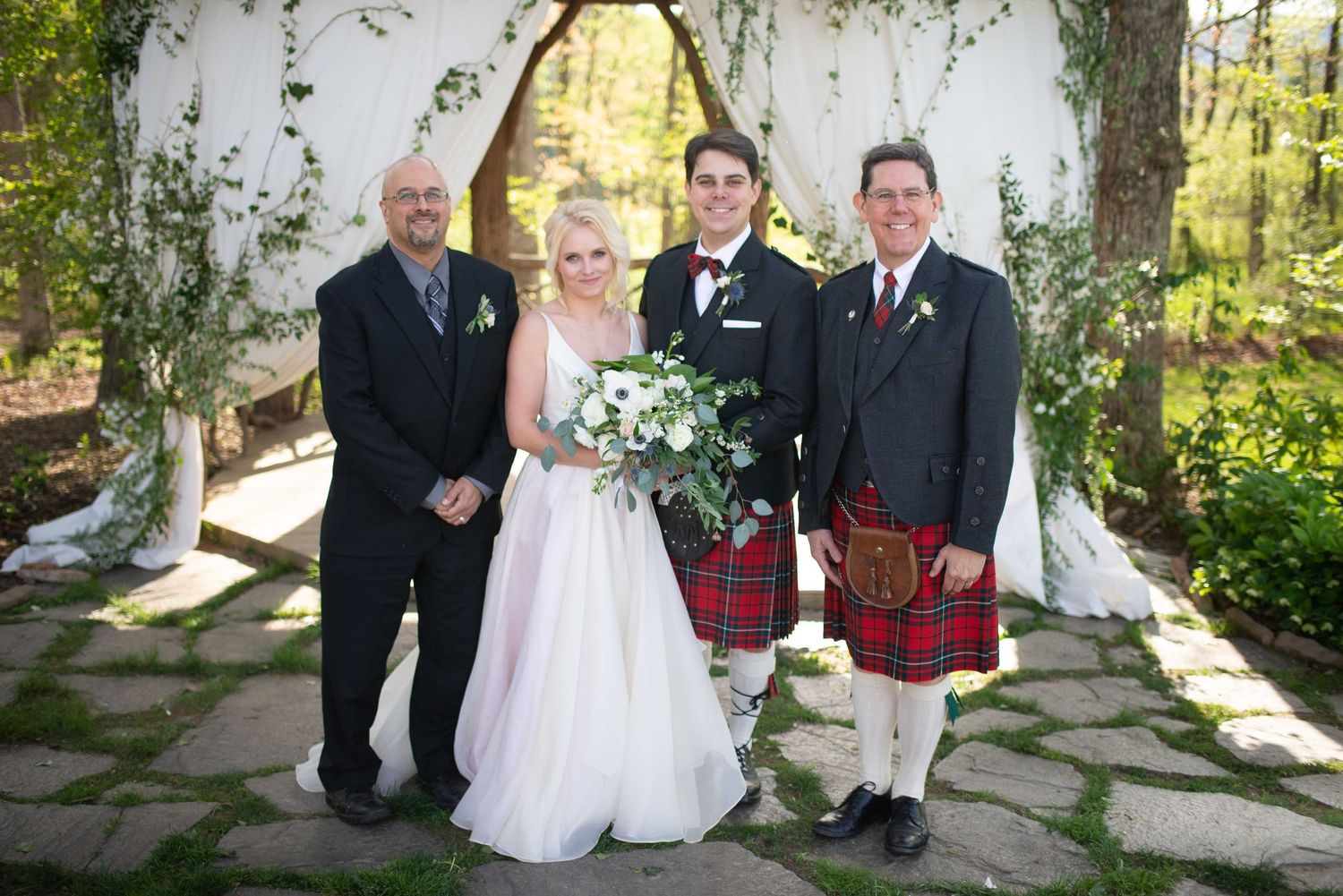 Wedding party poses outdoors under floral arch, bride in white gown with bouquet, groom in bow tie, man in red tartan kilt.