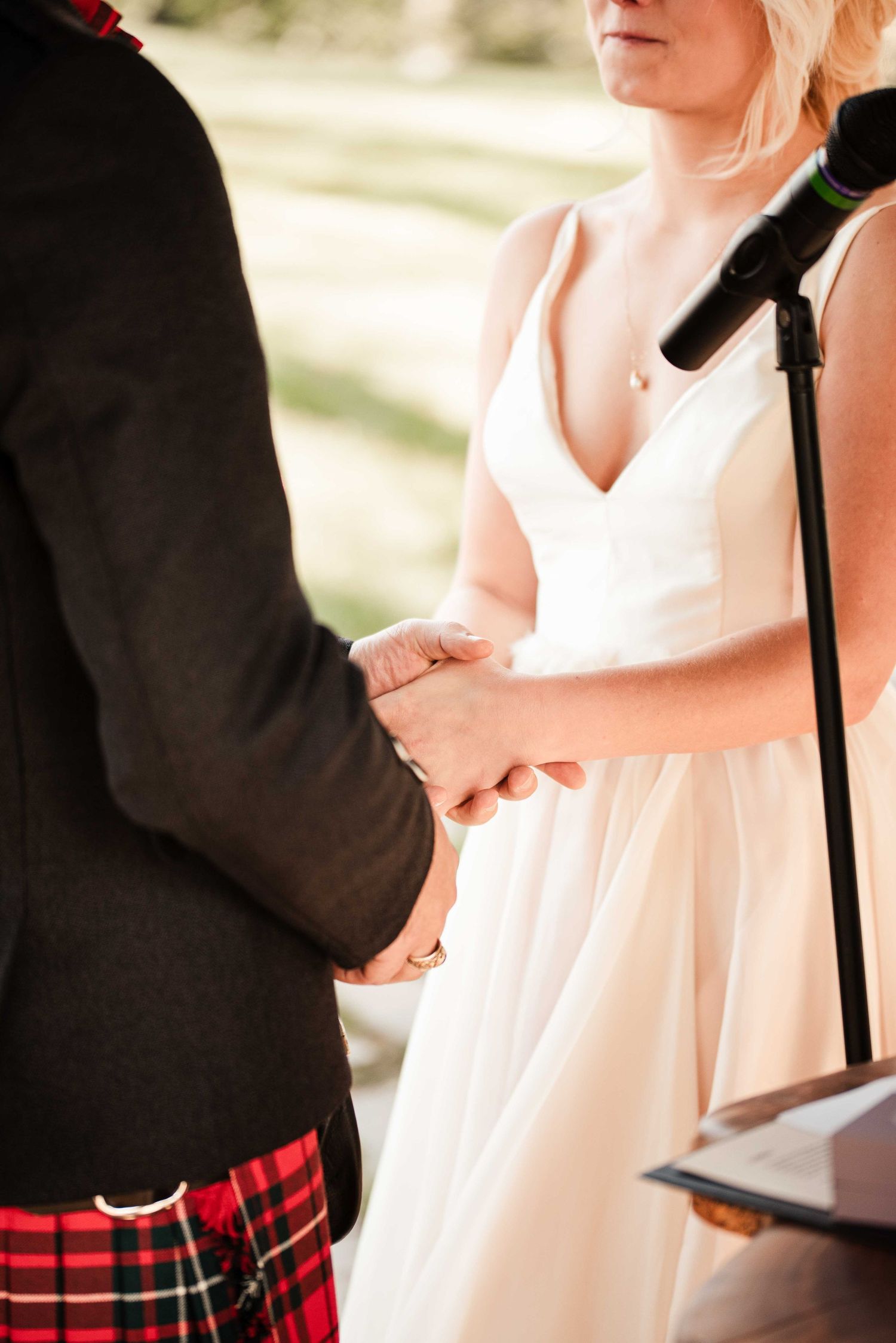 Bride in white gown holds hands with groom in red tartan kilt during outdoor wedding ceremony.