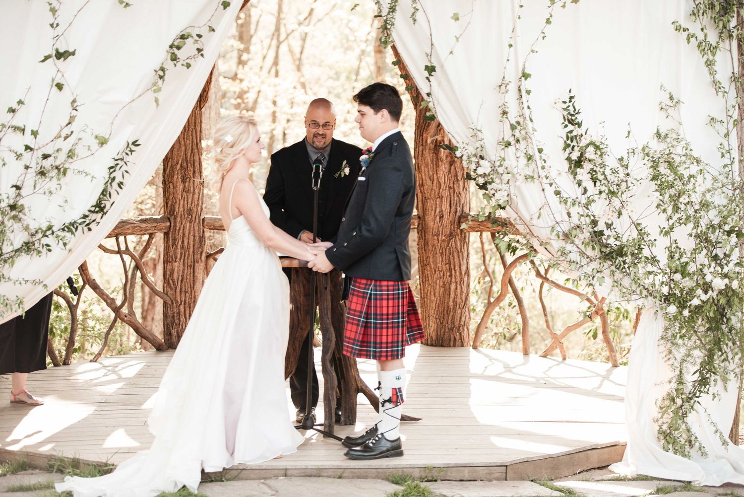 Bride and groom exchange vows under a rustic floral arch, groom wearing a traditional red tartan kilt at outdoor wedding.