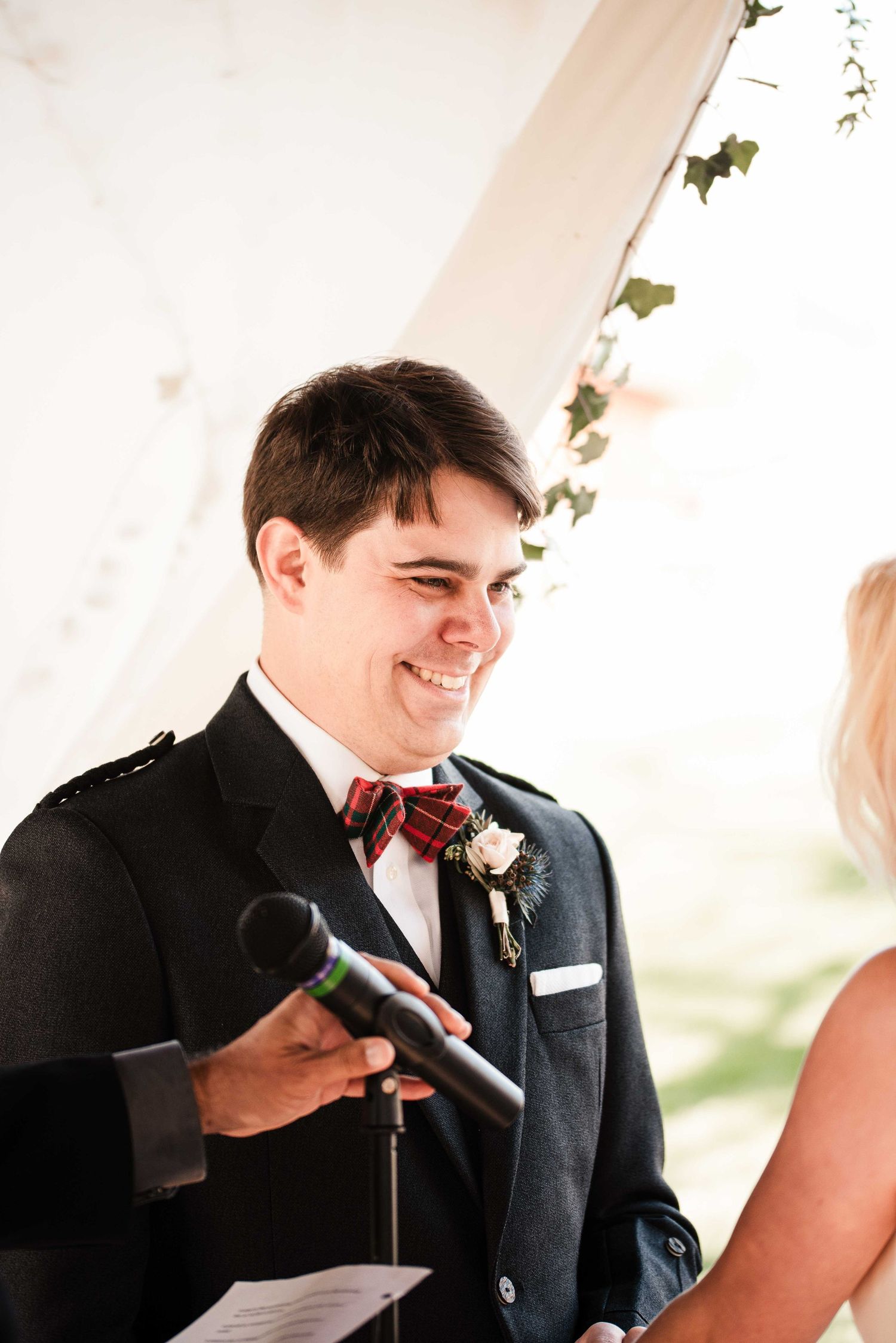 Smiling groom in dark suit with plaid bow tie and floral boutonniere holding microphone during outdoor wedding ceremony.