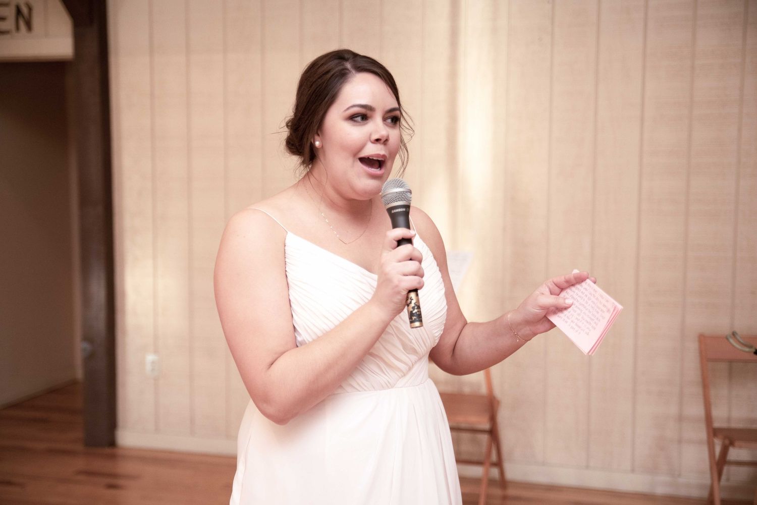 Woman in white bridesmaid dress speaks into microphone holding pink notes during wedding reception toast.