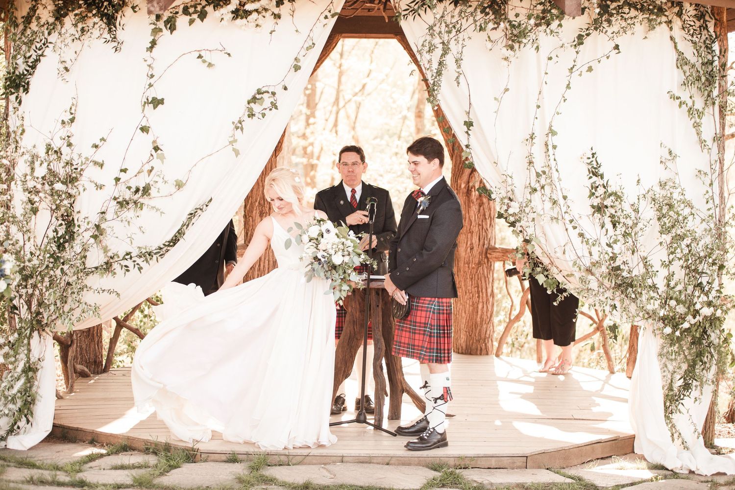 Bride and groom exchanging vows under white draped arch with greenery at sunny outdoor wedding ceremony.