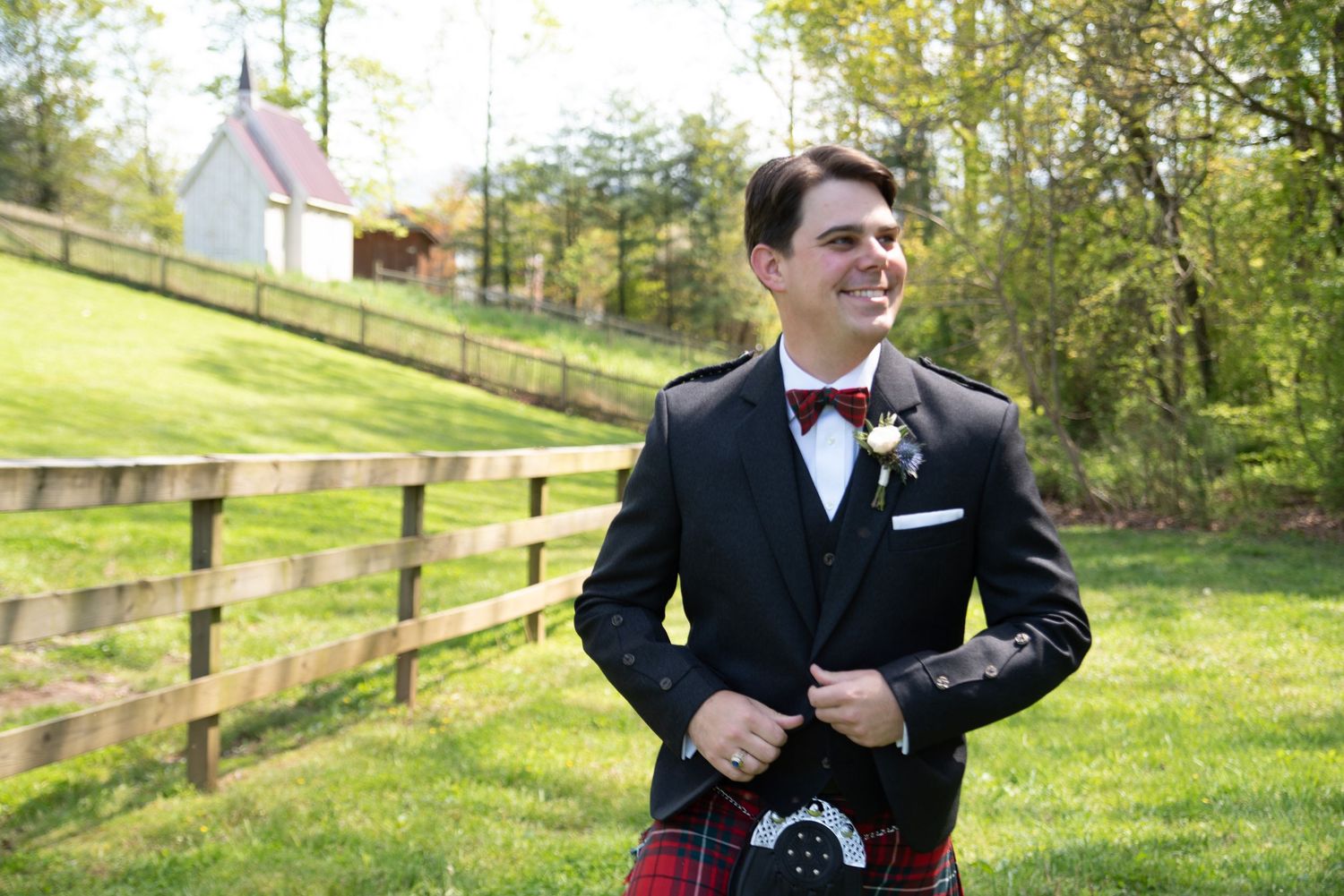 Handsome groom in black jacket, red tartan kilt and bow tie stands by wooden fence at outdoor wedding.