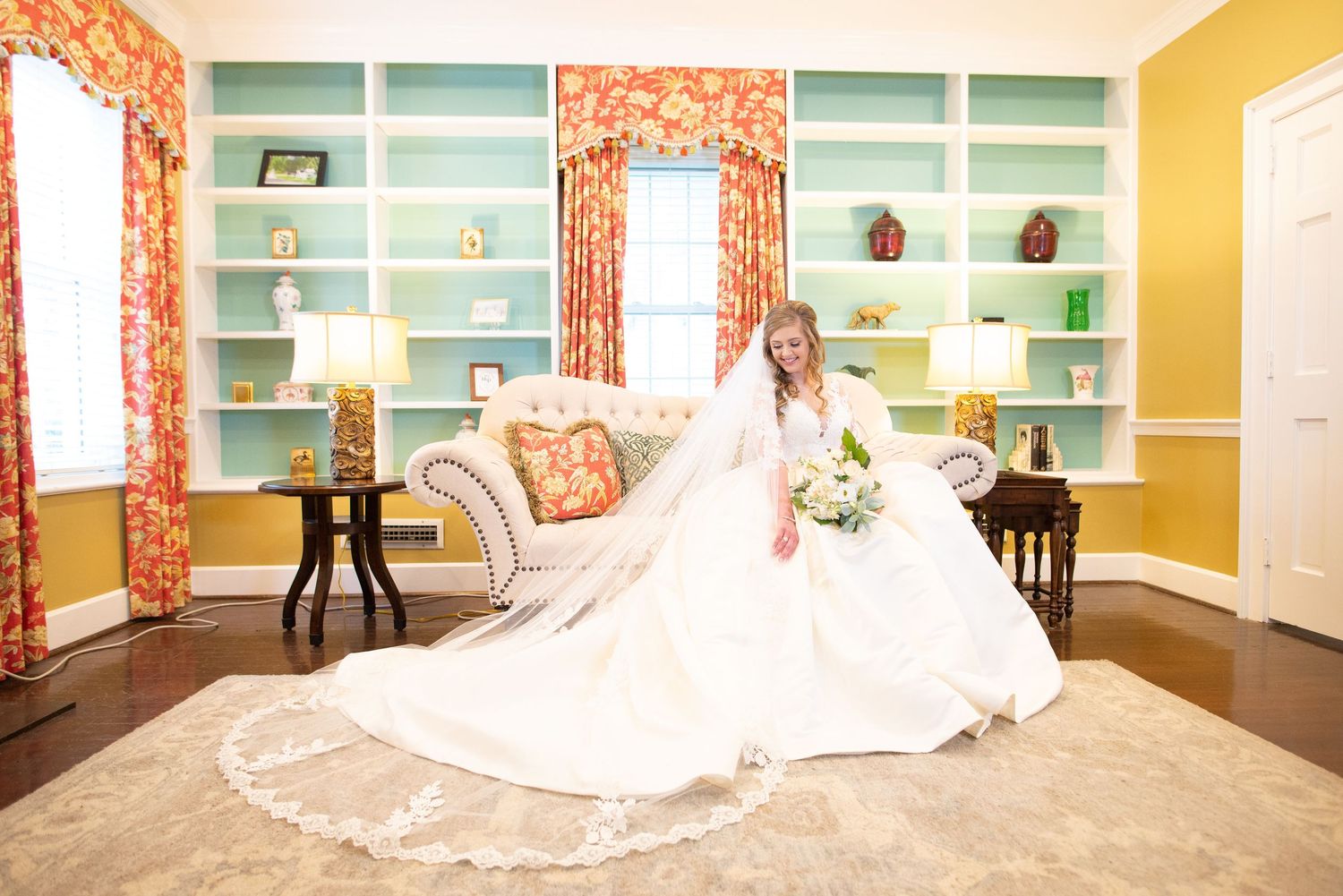 Bride in full white gown with long veil seated elegantly in colorful decorated room.