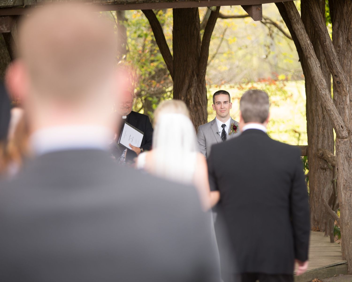 Groom awaiting bride during outdoor wedding ceremony under tree canopy with wedding party.