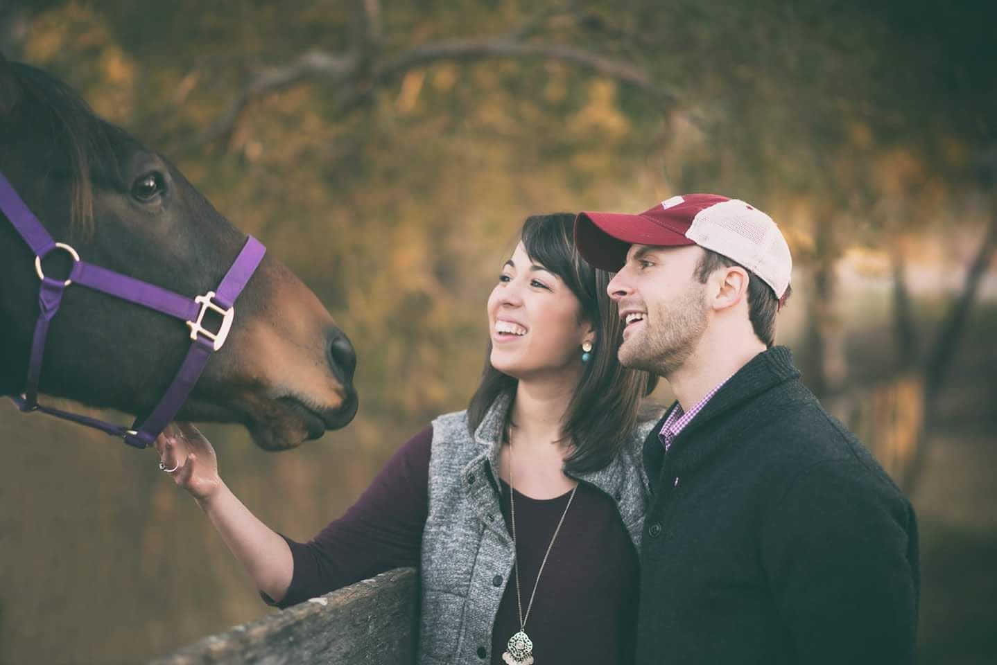 Smiling couple feeds and pets brown horse at rustic farm during engagement photo session.