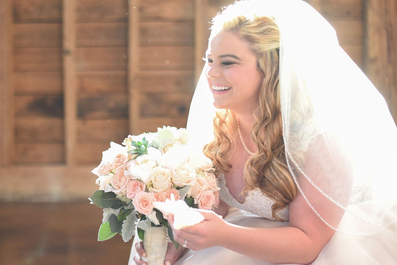 Smiling bride in white gown and veil holding blush and cream rose bouquet near rustic wood wall.