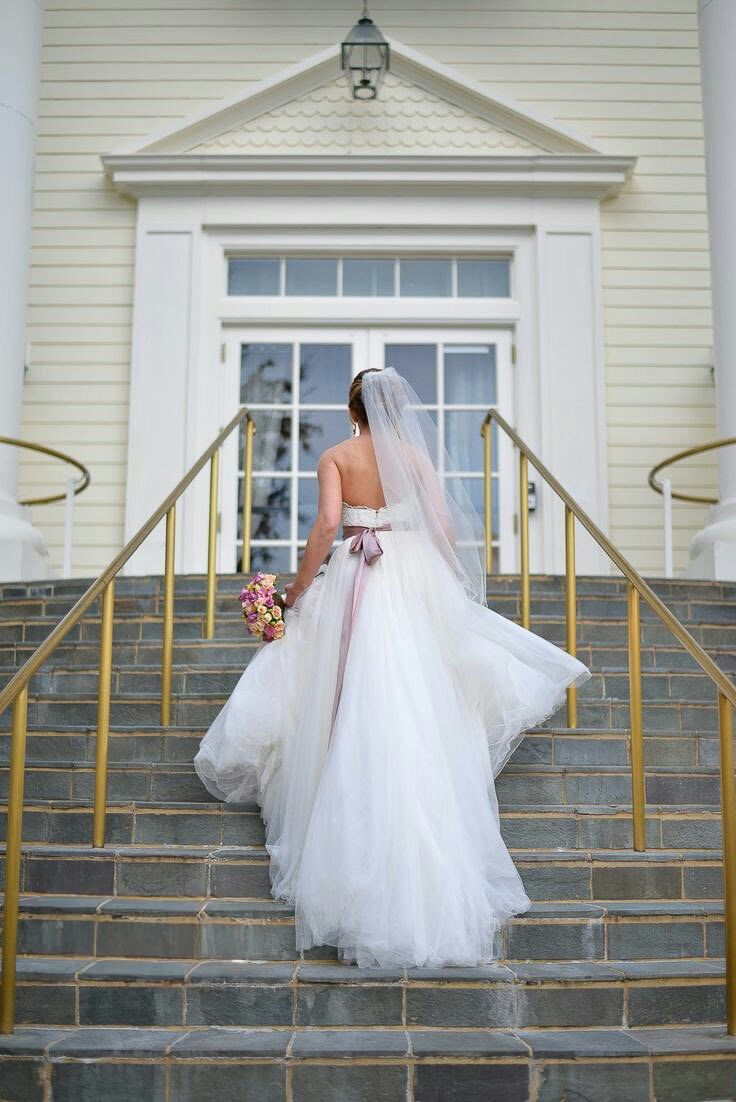 Bride in flowing white tulle gown with veil and colorful bouquet ascending stone steps of white church.
