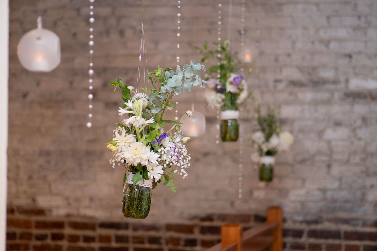 Mason jars with white and purple wildflowers hanging from crystal strands in a rustic brick wedding venue.