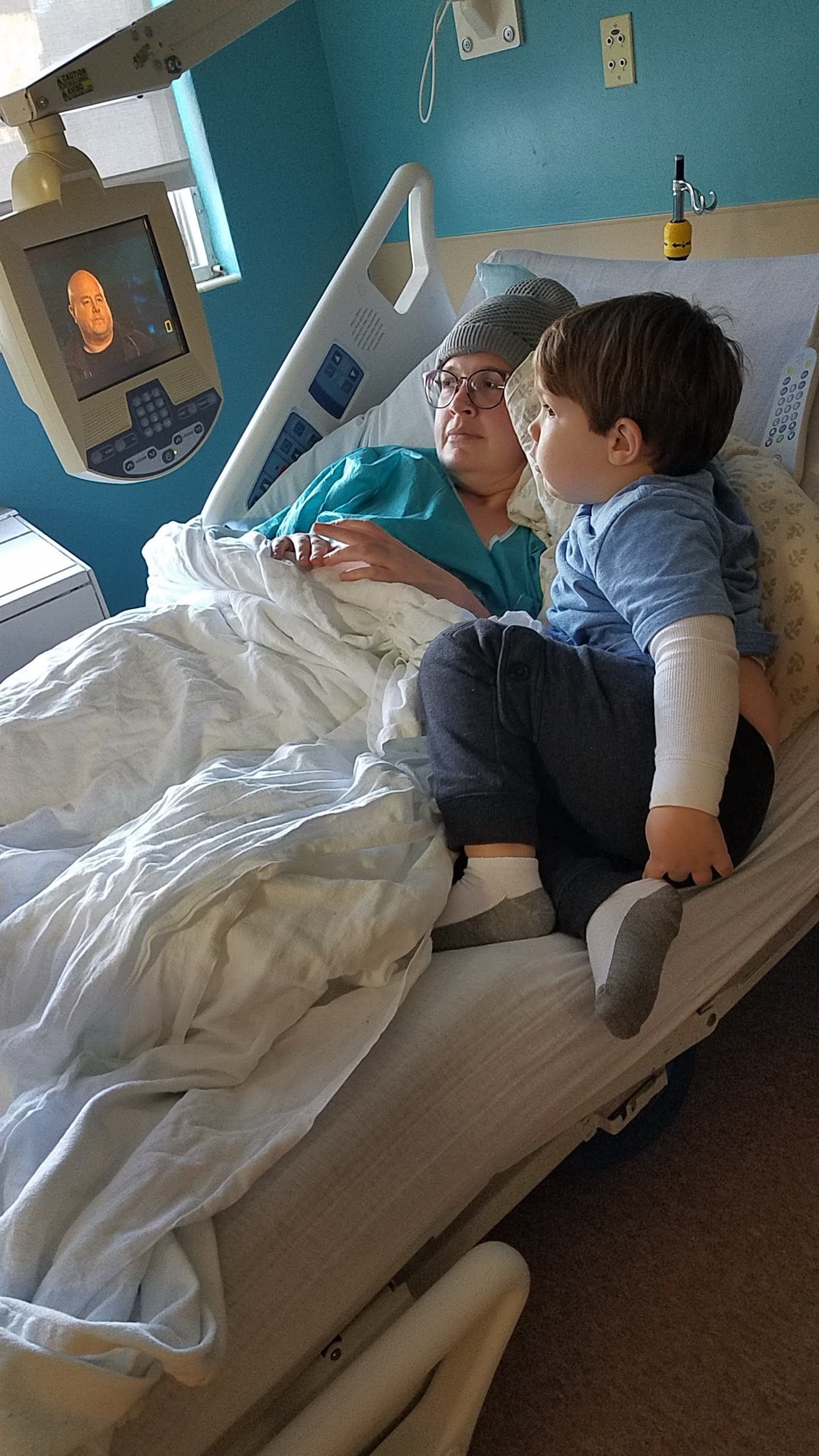 A woman in a hospital bed wearing glasses and a teal gown rests with a young toddler boy sitting beside her.