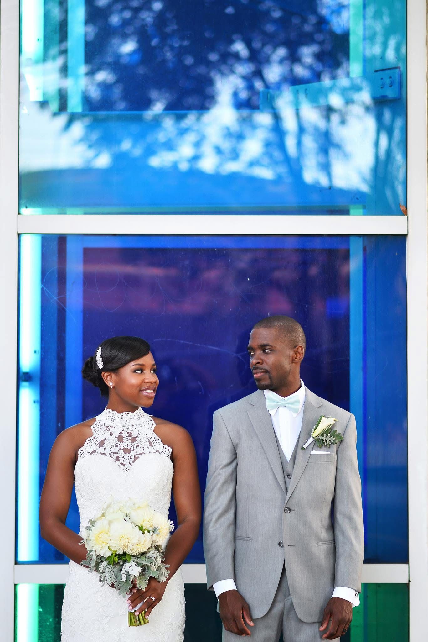 Bride in lace halter gown and groom in gray suit posing before vibrant blue glass doors.