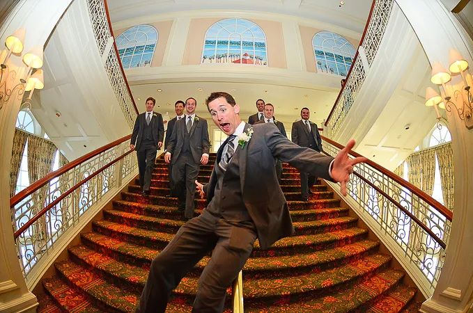 Groom poses playfully on grand staircase with groomsmen behind him in fisheye lens photo.