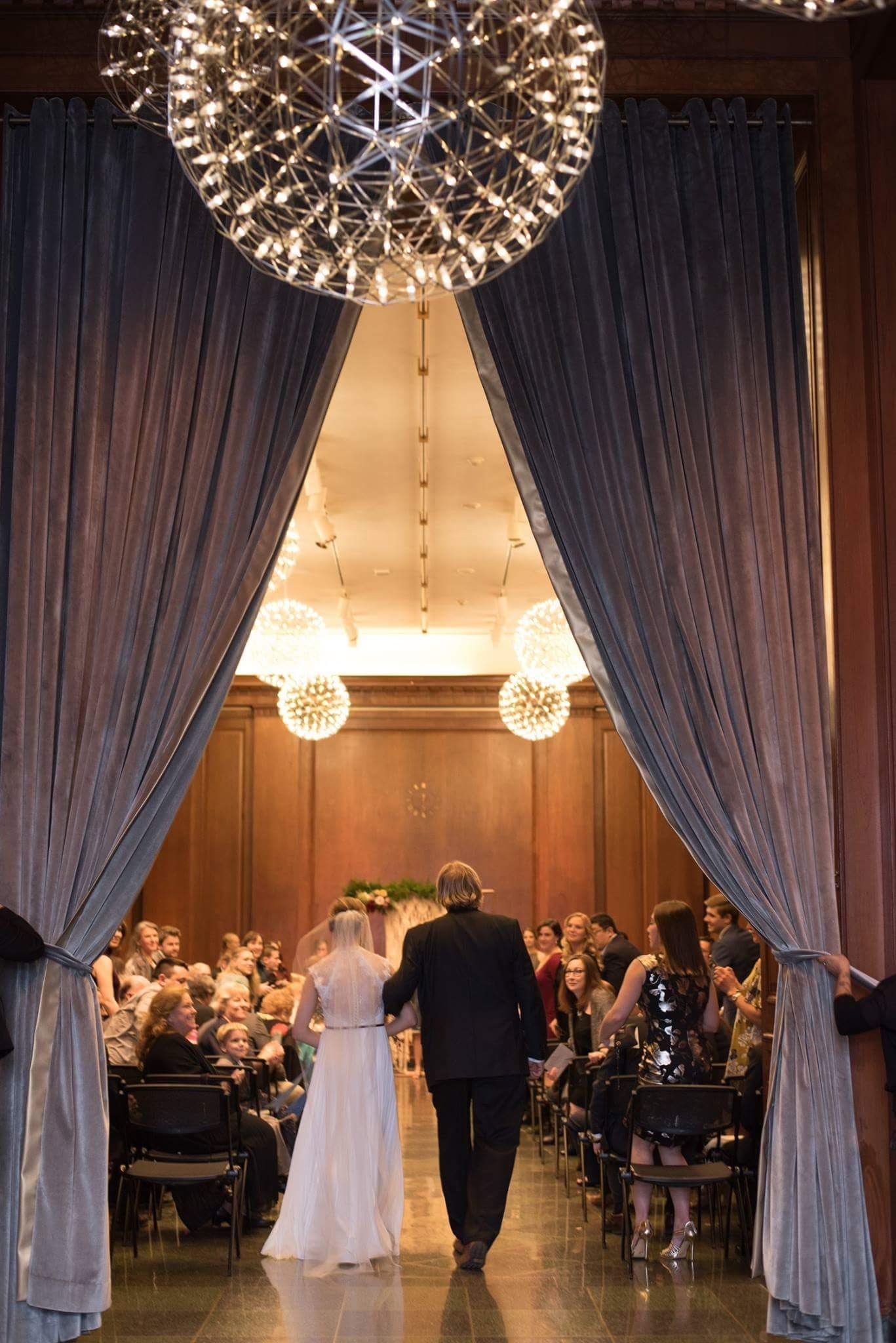 Bride and escort walk down the aisle through dramatic draped curtains under sparkling globe chandeliers indoors.