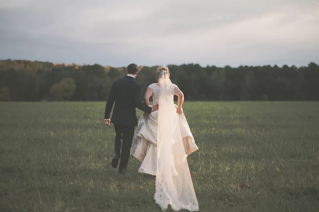Bride and groom walking hand in hand through a lush green field with a forest backdrop on their wedding day.