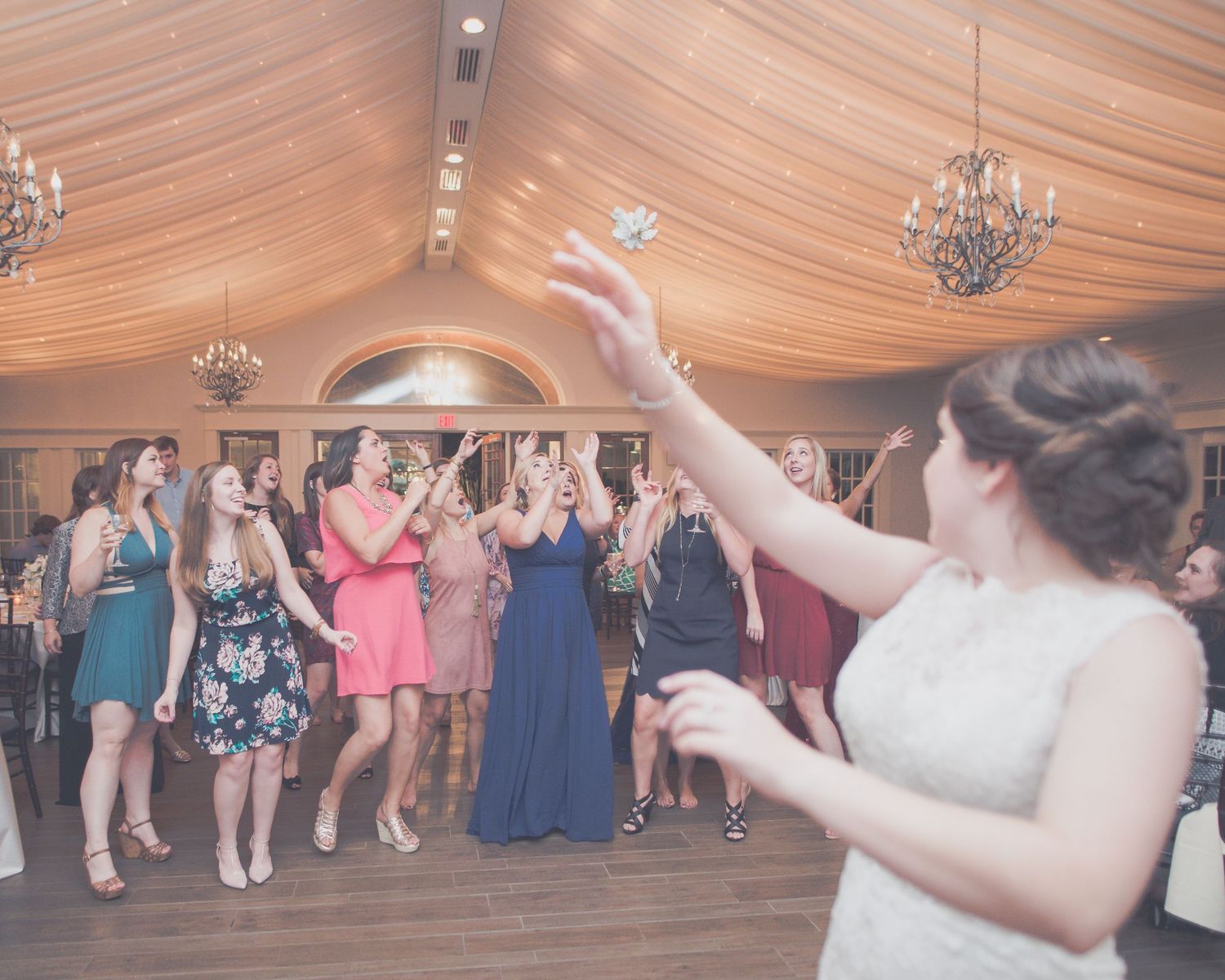 Bride tossing bouquet to excited guests in decorated reception hall with draped ceiling and chandeliers.