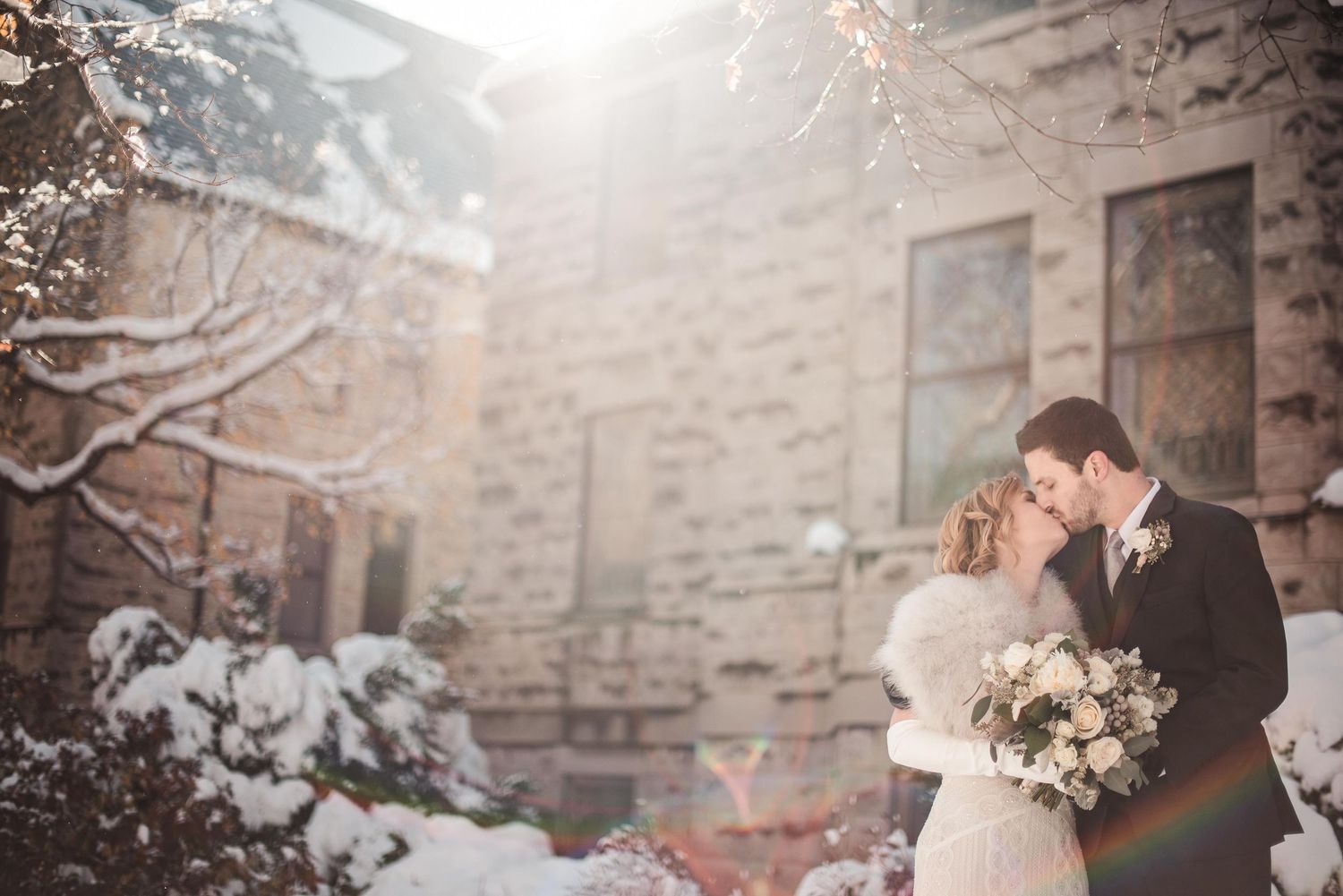 A bride and groom share a romantic kiss outdoors in a snowy winter wonderland, surrounded by snow-covered trees and a stone building.