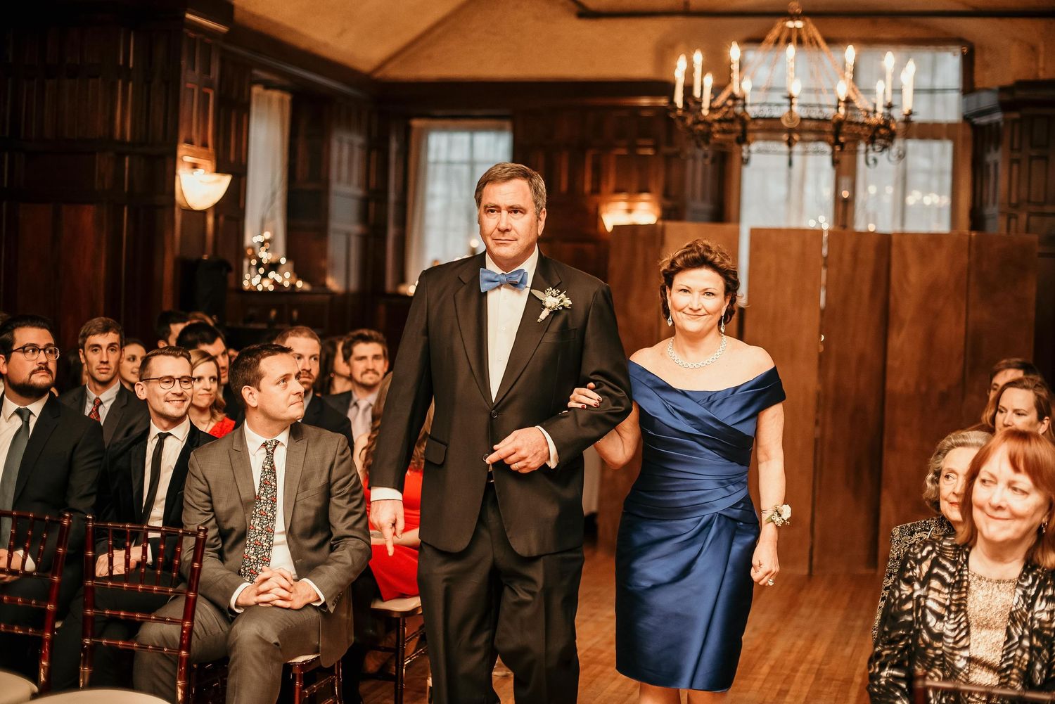 Groom in tuxedo walks down wedding aisle with woman in blue dress in elegant wood-paneled venue.