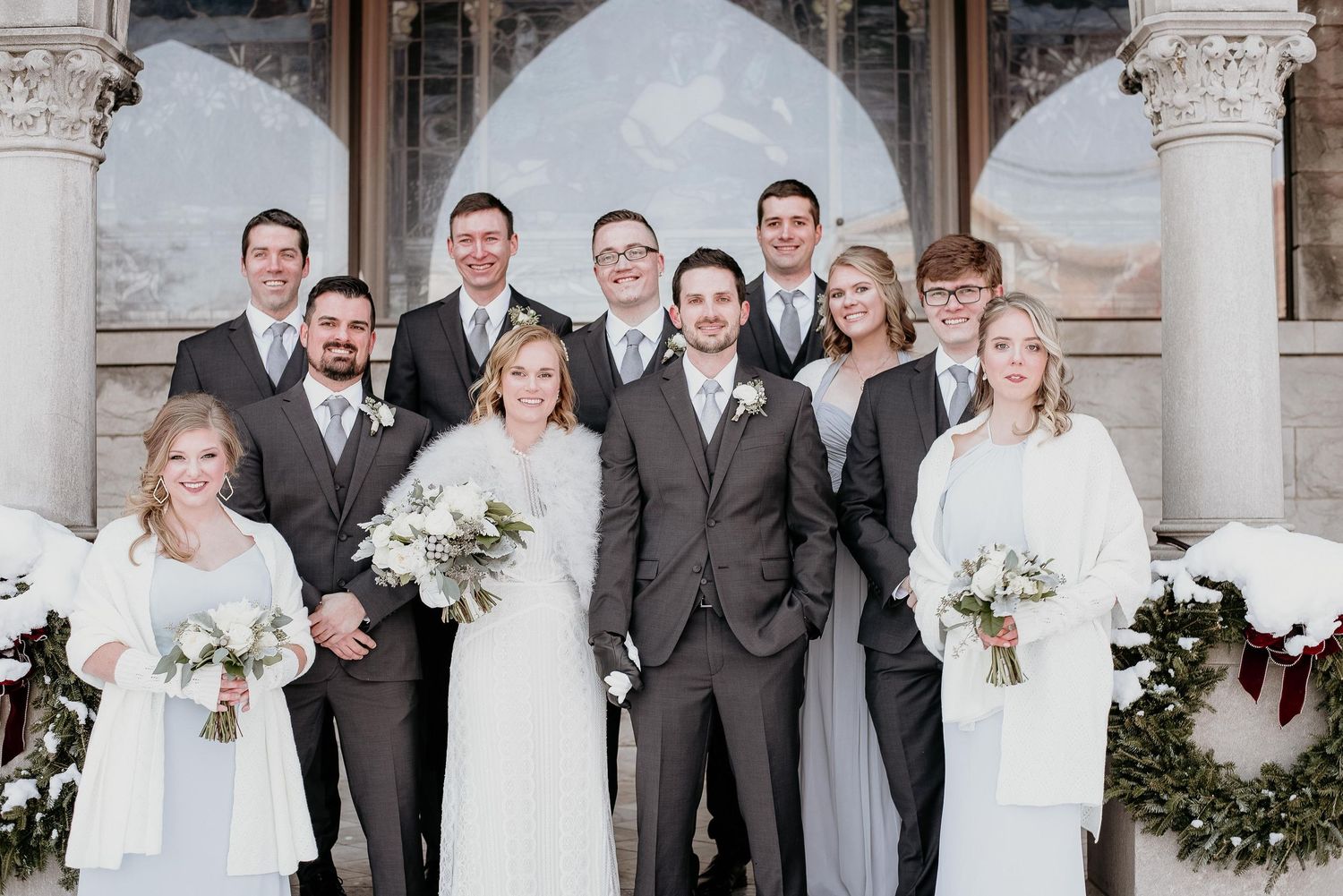 Wedding party poses in gray suits and white dresses with bouquets outside an elegant venue in winter.