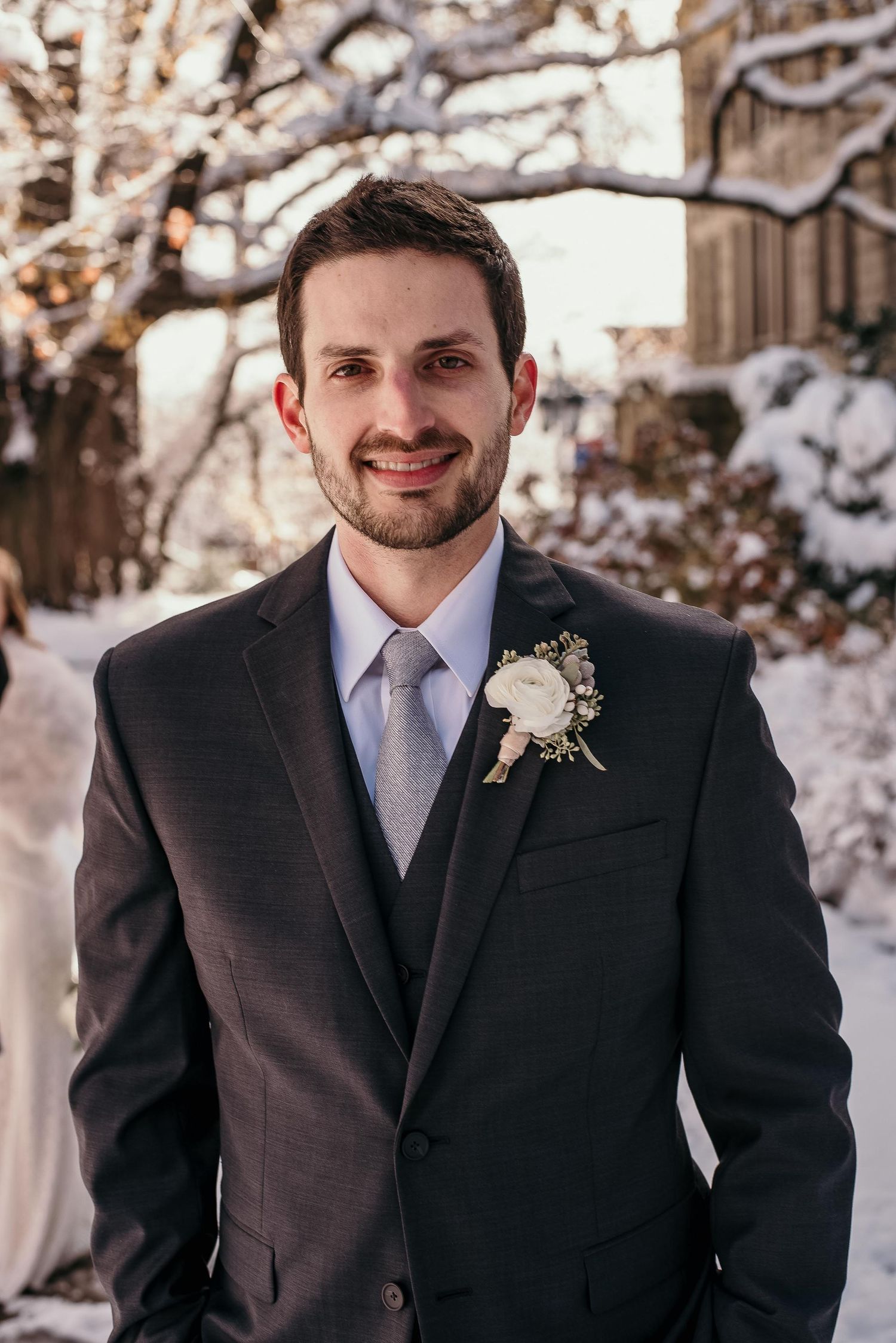 Smiling groom in dark suit with boutonniere and gray tie poses outdoors in snowy winter wedding setting.