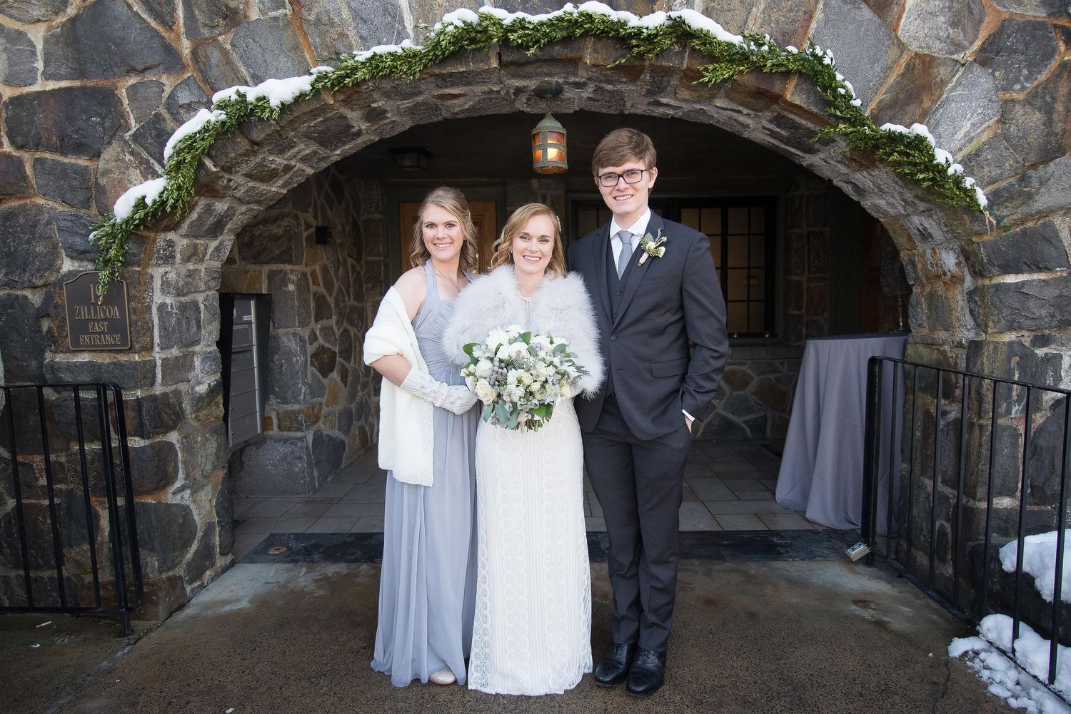 Bride with bouquet and two guests posing under stone arch at winter wedding venue with snow-dusted greenery.
