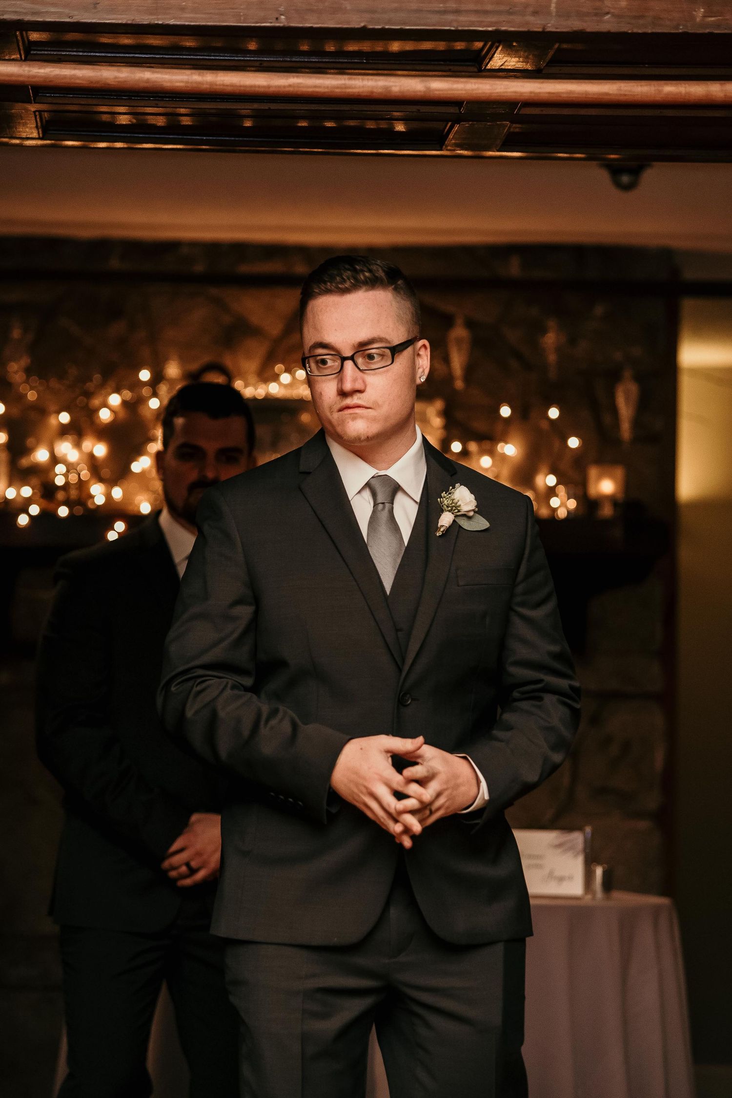 A young man in a dark suit with boutonniere and glasses stands at a wedding reception with warm string lights behind him.