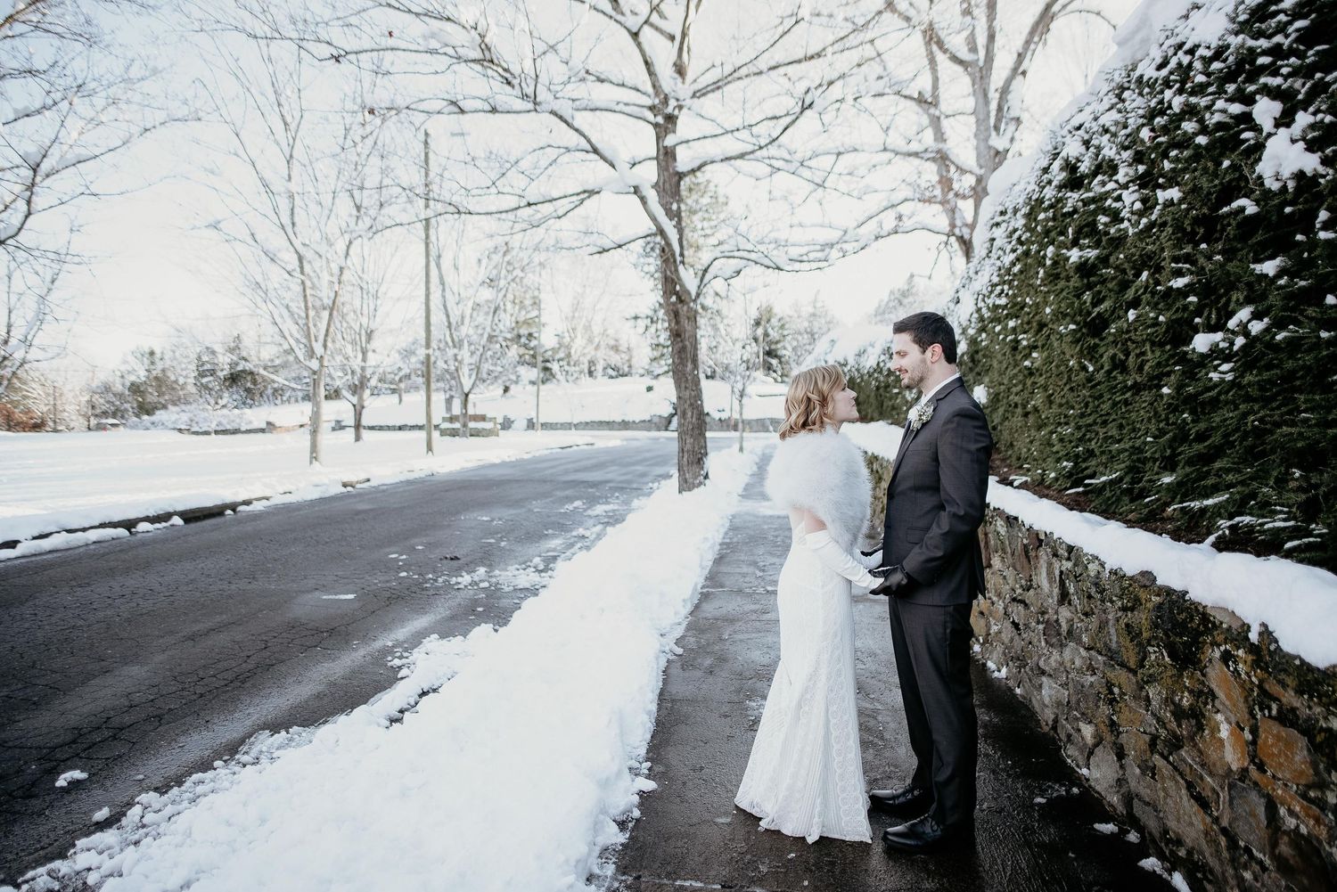 Bride and groom share a romantic moment on a snowy winter wedding day beside a stone wall.
