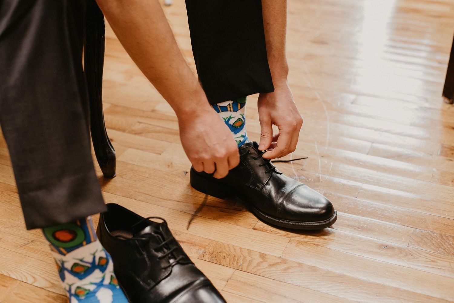 Man tying black dress shoes while wearing colorful patterned socks and gray trousers on a wooden floor.