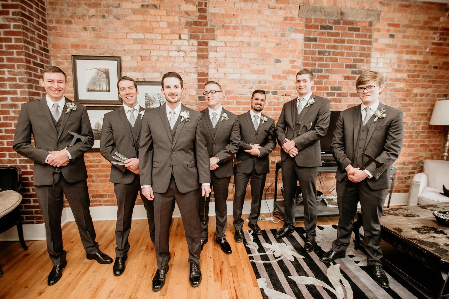 Groomsmen in matching gray suits holding shoes, posing together in a rustic brick-walled venue with hardwood floors.