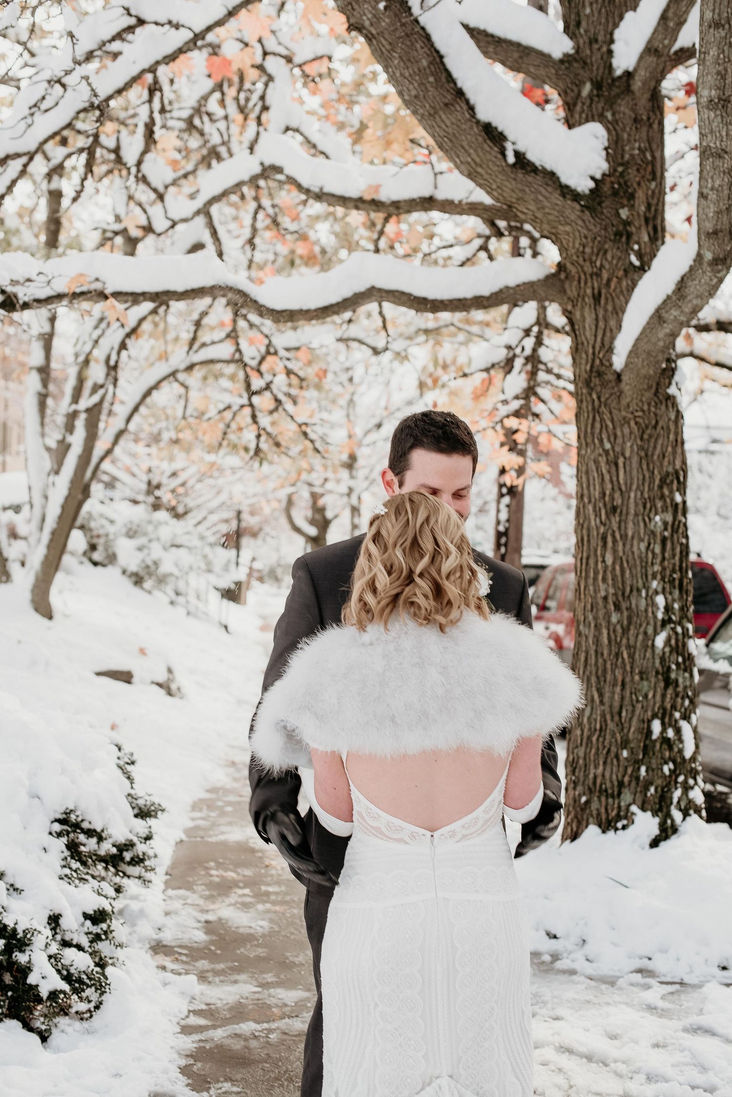 Bride in white lace gown with fur shawl embraced by groom in snowy winter wedding outdoor setting.