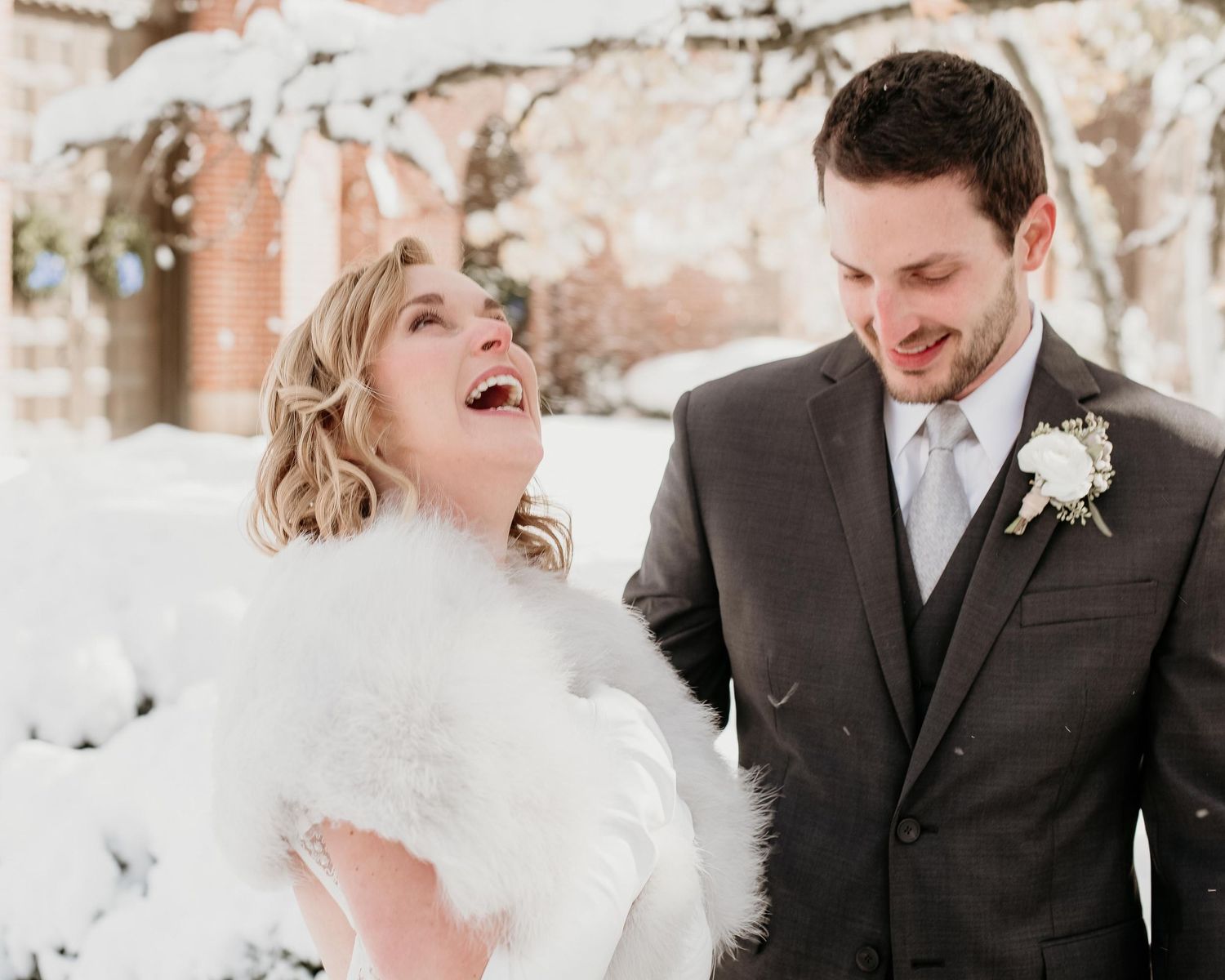 Happy bride in white fur wrap laughing with groom in gray suit during a snowy winter wedding outdoor photo.