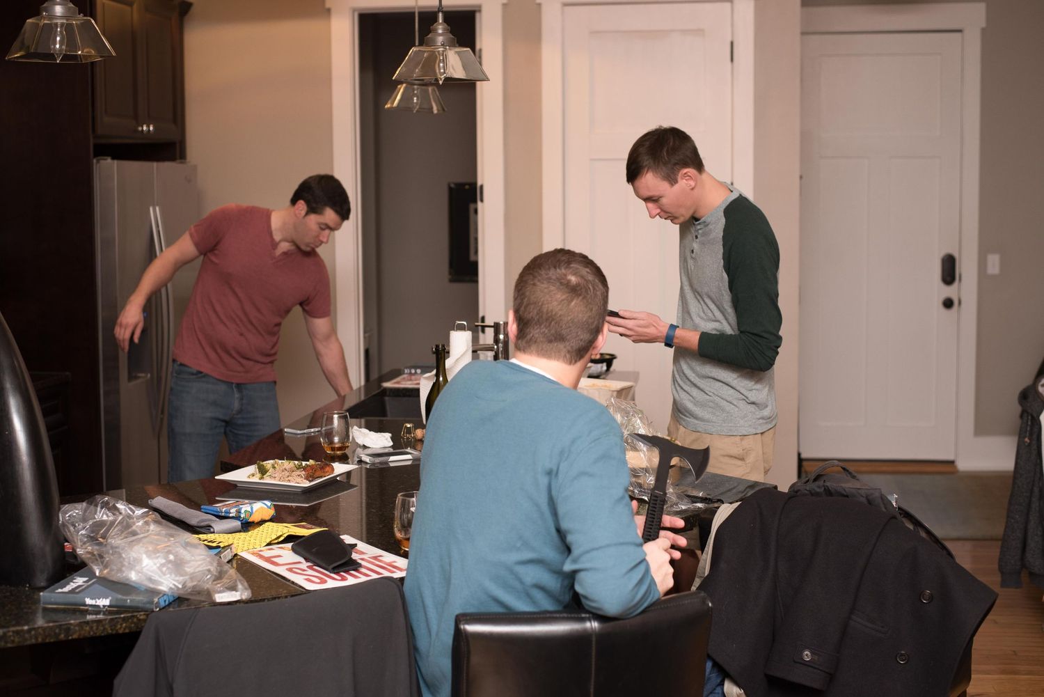 Three men gathered in a modern kitchen and dining area, with one cooking, one standing, and one seated at a table.