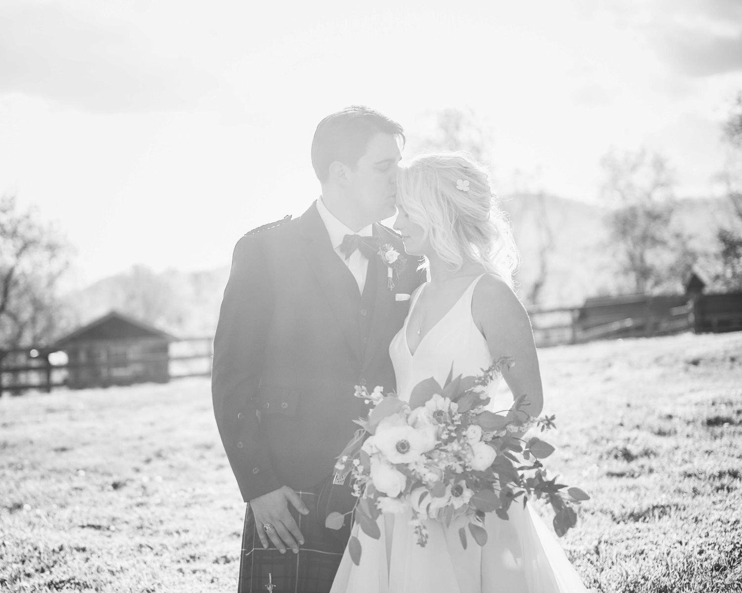 Black and white photo of a bride and groom sharing a romantic moment in a sunlit field with a rustic fence backdrop.
