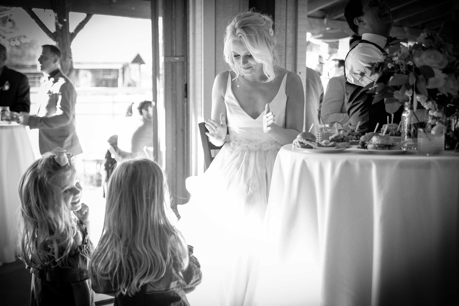 Bride in white gown interacts with two young flower girls at a wedding reception table in black and white photo.