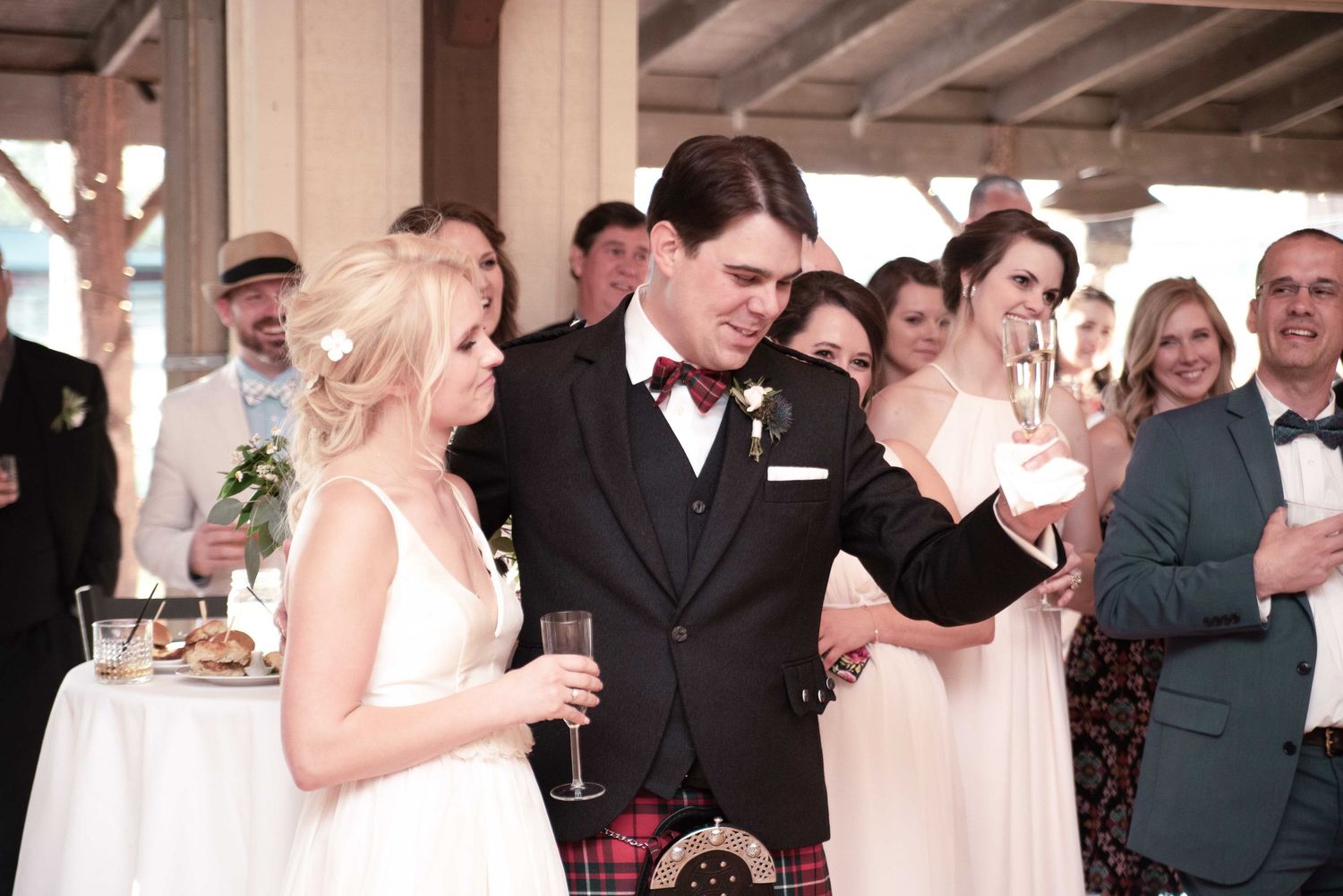 Bride and groom in tartan kilt share a toast with champagne at their wedding reception surrounded by guests.