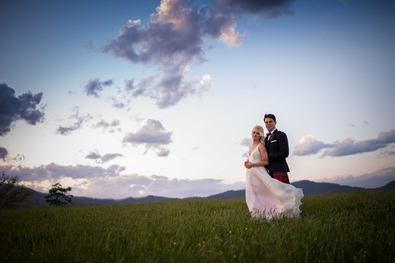 Bride and groom standing in a lush green field at sunset with dramatic clouds and mountains in the background.
