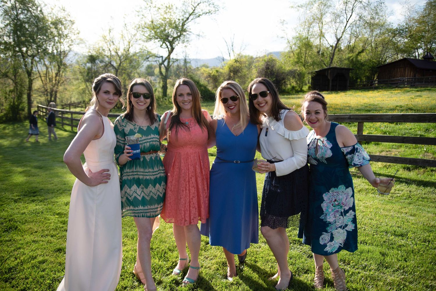 Six women smiling and posing together outdoors on a sunny day at a rustic farm venue.