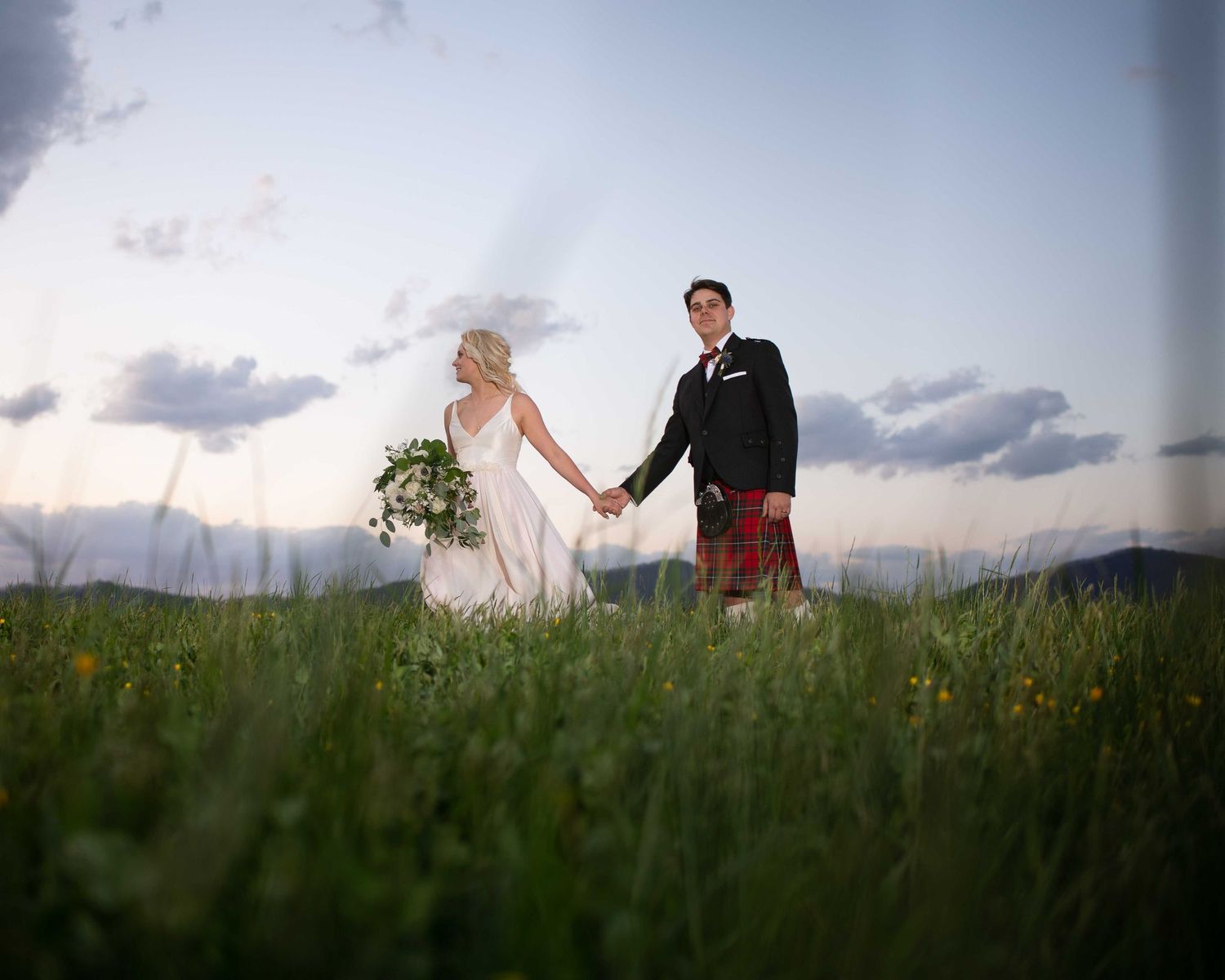 Wedding couple holding hands in lush green meadow at dusk, groom in red tartan kilt, rolling hills behind them.
